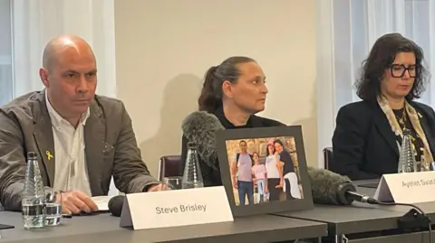 Pictured sitting at news conference, Steve Brisley, Ayelet Svatitzky, and Sharone Lifschitz all have serious expressions. On the table in front of them are bottles of water and their name tags, with a picture showing one of their families