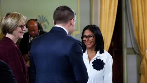 REUTERS/Leonardo Fernandez Viloria Venezuela's interim president Delcy Rodríguez meets Laura Dogu, U.S. Charge d'Affaires to Venezuela and U.S. Assistant Secretary of Energy Kyle Haustveit during a meeting with oil executives, in Caracas, on 13 April, 2026.
Rodríguez is smiling. She is wearing black-rimmed chunky glasses and a white blouse adorneed with a black decoration on her chest. Laura Dogu smiles as she greets Rodríguez. Haustveit is wearing a navy suit and has his back to the camara. 