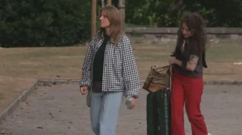 RTÉ Image of Markéta Irglová with brown hair, and a black and white jacket. She's wearing blue jeans. Beside her is a woman with dark hair, black shirt and red trousers. She is carrying a suitcase.