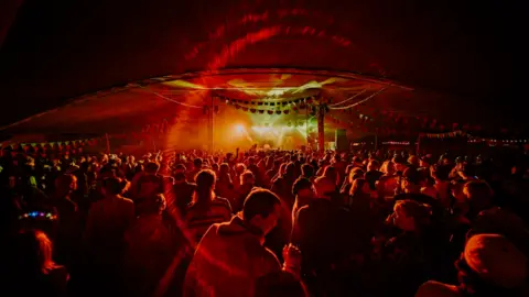 Shindig Festival Crowds of people at an event waving their hands and dancing in front of a stage illuminated by spotlights at the Shindig Festival