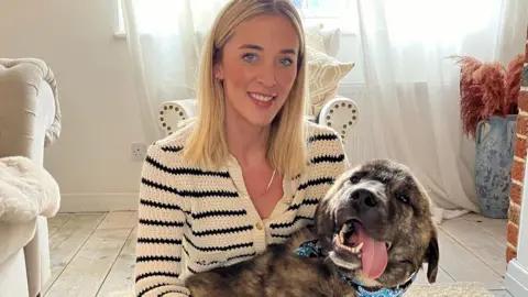 A dog with a cloth round his neck and his tongue out in the embrace of his foster mum who's wearing a striped top and is smiling at the camera