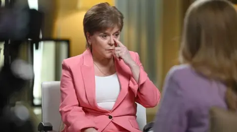 Nicola Sturgeon, wearing a pink suit, touches her nose as she is being interviewed