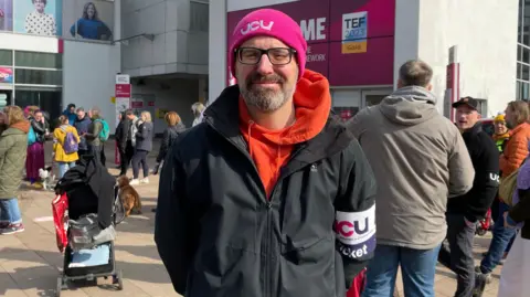 A man with a greying beard and wearing glasses and a pink beanie hat with a UCU logo is standing facing the camera in front of several other people on a picket line and at the entrance to Sheffield Hallam University's Owen Building.