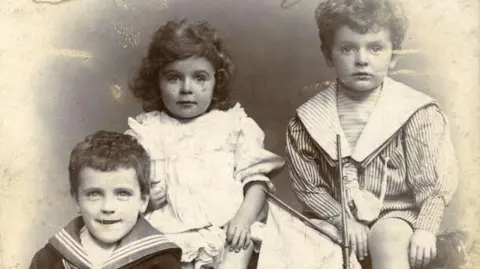 Leeds City Council A Victorian, sepia photograph showing three young children wearing fashions from the era, including sailor suits.