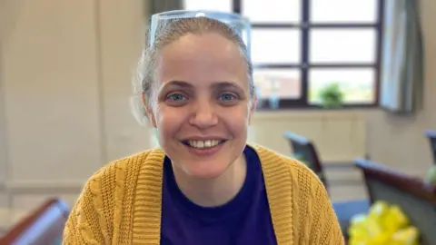 A women with a blue t-shirt and yellow jumper. She has curly brown hair which is tied back and clear glasses on her head. She is looking at the camera smiling.