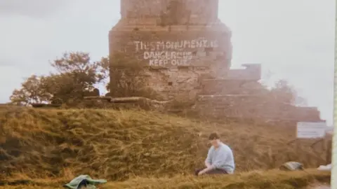 Gary Haines A woman sat on a grassy hill in front of a brick monument, it is an old photograph with a sepia tone