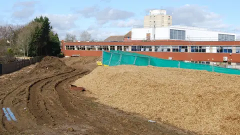 Enabling works being carried out on the extension to Kettering General Hospital's maternity unit, showing mud on the ground, green barriers, a building behind it and stone on the ground. 