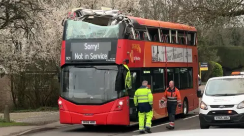 A red and orange double-decker bus is pictured stopped in the road. The upper deck is significantly damaged following a crash with a bridge. A message reading "Sorry! Not in Service" is being shown on the bus