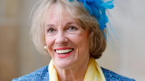 Dame Esther Rantzen, an elderly woman with a short blonde bob, wears a blue feathered fascinator, a blue jacket and a yellow scarf
