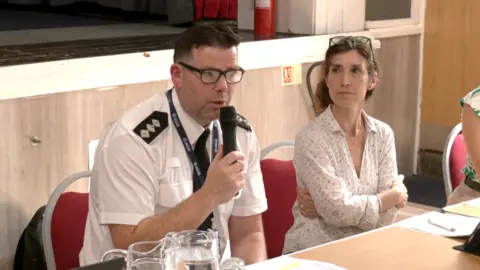 A man with a white shirt and black tie holds a microphone. A woman wearing a white shirt sits next to him with her arms crossed. Both are sat at a table. 