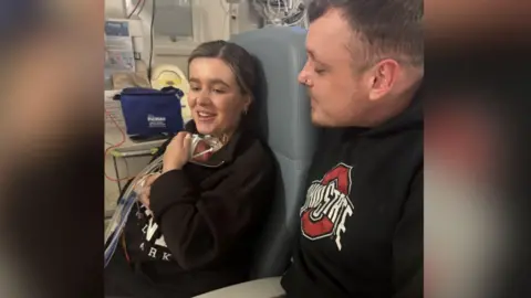 Family handout Chelsea Gowar holds her baby Bonnie close to her chest while sitting on a hospital chair in a maternity department while her partner sits alongside her looking at the baby.