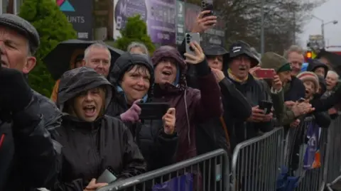 Jamie Niblock/BBC Crowd of people all wearing rain coats with hoods up stand behind a barrier cheering. 
