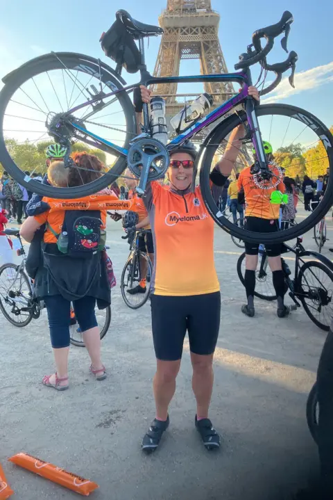 Deb Gascoyne The same woman, wearing black lycra shorts and wearing a similar lycra top to the above. She is also wearing sunglasses and a cycling helmet as she holds a bike up in the air with the Eiffel Tower in the background as other people with bikes mill around.