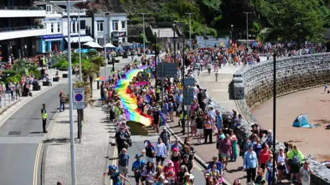 Harry Schofield A crowd of people carrying a large pride flag walking along a road. To the left are houses and two the right is a beach. 