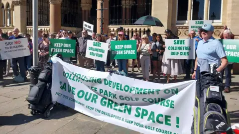 Lots of people stand in front of a stone building. They are holding green and white placards with "Keep our space" and "Save Collingtree Golf Club" written on them. There is also a large white banner bookended by two golf club bags with "Housing development on Collingtree Park Golf Course. Save our green space," written on it. It is a sunny day.