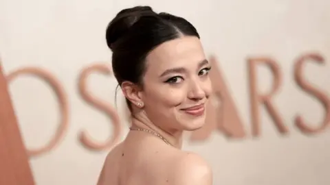 Best Actress winner Mikey Madison looks back over her shoulder and smiles at the camera, in the Oscars press room. Her dark hair is tied up and she wears a necklace.