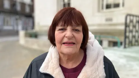 Sam Read/BBC Hazel Simmons with dark red hair in a bob, looking at the camera and wearing a black coat with white trim, and a red sweater. She is standing in front of the stone-built Town Hall