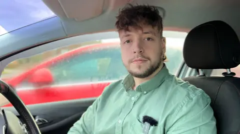 A man with a mop of dark-brown hair and a smart beard is sitting in the driving seat of a car. He is wearing a green shirt and looking towards the camera.