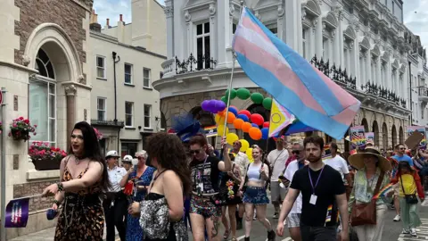 The photo shows a vibrant parade on a city street with participants in colourful attire, some holding balloons and flags. One person prominently carries a large transgender Pride flag. The background includes historic buildings with ornate architecture, flower boxes on the windowsills, and a blue sky.