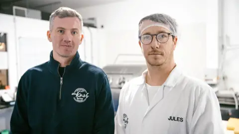 Kieran Woods is wearing a blue fleece with the Knob of Butter logo on it. He is standing next to Jules Bal who is wearing a hair net, glasses and a white T-shirt with a white overall over it. It has the name Jules sewn into it.