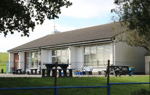 Billy McCrorie A small school building with a low tiled roof and a white frontage surrounded by picnic tables, grass and a low fence