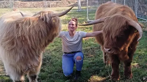 Chloe Lucas Chloe Lucas kneeling on grass with two of her cows next to her. Both have brown fur, although one is slightly darker than the other. Chloe has one arm on each cow and is smiling.