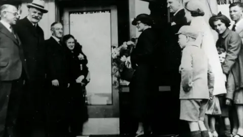 KELLANOVA Black and white photograph showing the opening of the plant with a crowd of people and a woman with flowers in the foreground.