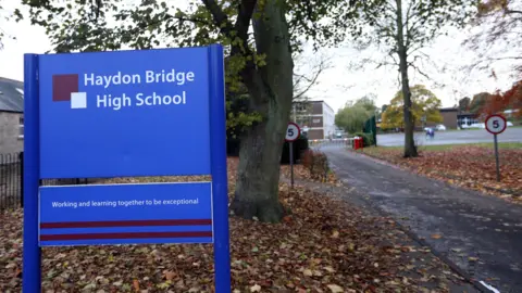 A blue sign that reads in white lettering, 'Haydon Bridge High School' on a patch of grass covered in brown leaves. It stands in front of a tree. To the right is a path leading to a school. 
