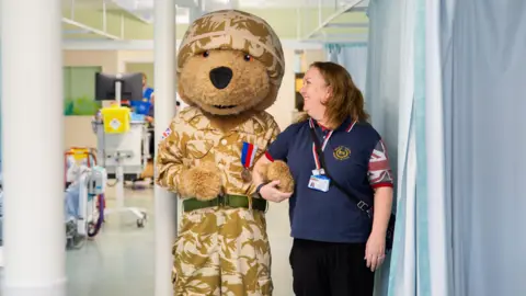 Cat Cusack A Help for Heroes teddy mascot dressed in camo army gear. Julie Wadham is standing next to the mascot, linking arms with him. She has her head turned to the side, looking at the mascot. She is smiling. She is wearing a navy blue Help for Heroes polo shirt which has union jack prints on the sleeves. They are standing in a hospital ward - blue curtains, beds and hospital machines can be seen in the background.