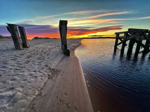 Douglas T Coutts Landscape image of a beach with a vivid pink, purple, orange and yellow sunrise on a light blue sky with wooden pillars from an old pier on the beach