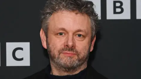Michael Sheen, dark hair and beard, staring straight-faced at the camera