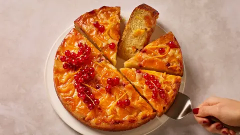 A yellow cake with berries on top, with half cut into slices, on a pink background.