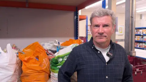 David Holt, a man with dark hair, looks at the camera. He’s wearing a dark blue checked shirt and stands in front of Foodbank bags.