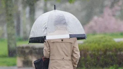 PA Media A woman walks in the rain, she has a beige coat and black handbag, and holds a see-through umbrella.