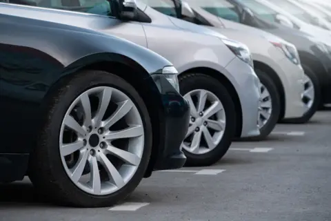 A closeup of the fronts of several cars parked pointing to the right of the image. The cars are a mixture of black, silver and white.