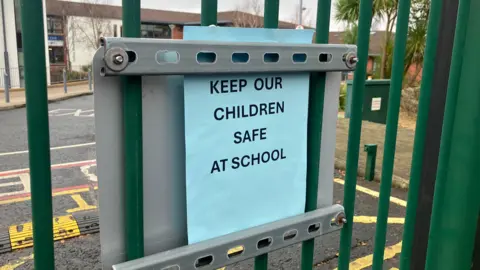 On the green metal gates is a blue sign that reads 'Keep our children safe at school'.