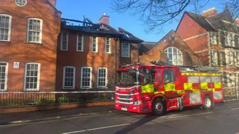 BBC A fire engine outside a red-brick building with large sash windows and white window sills. A large part of the roof has been completely destroyed by fire, with wooden beams visible from the road. 