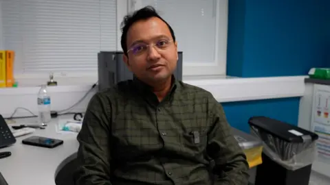 A man sits in an office chair at a desk in a clinical consulting room. He is wearing a dark green checked shirt and is positioned in front of a computer monitor, with white blinds covering the windows behind him. A desk phone, mobile phone, water bottle and paperwork sit on the desk, while a bin and storage trays are visible to the right. The room has bright blue walls and white worktops, giving it a clean, modern appearance.