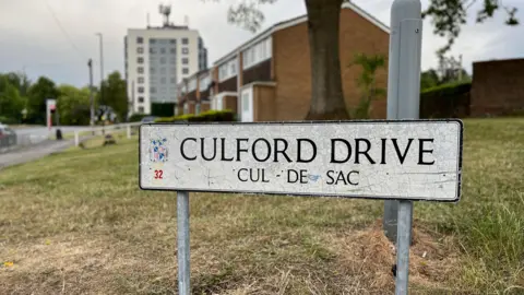 A street sign of Culford Drive, a cul-de-sac. It is old and scratched. The background is blurred but we can see some residential homes and a tower block behind it. 