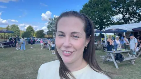Hayley James pictured with long brown hair and wearing a pale yellow top. She is stood in a large field with wooden benches and stalls behind her. Small groups of people are chatting and having a drink. 