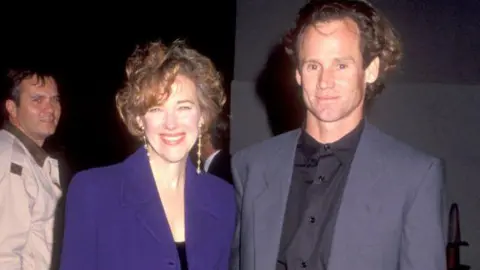 Getty Images O'Hara pictured smilng at an event in 1990 with a short brown bob, a purple blazer and black dress, with Bo Welch, who wears a grey suit and black shirt 