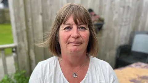 A woman in her 50s is sat on a garden bench with a fence behind her. She has mid length brown hair in a bob hair cut, and is wearing a white top with a silver necklace. She has a passive expression. 