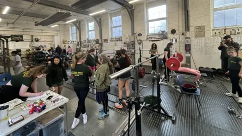 Inside of weightlifting gym: shows weights, people using it and black floor. A table is on the left