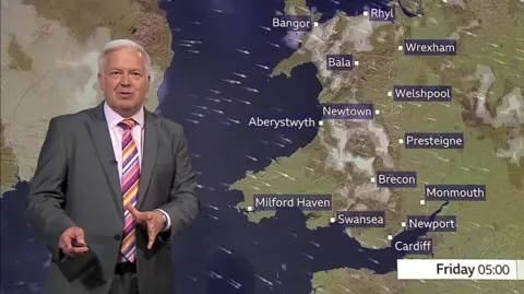 BBC Derek Brockway wearing a grey suit, a colourful tie and pink shirt. She is standing in front of a weather map showing Wales and various towns and cities. 