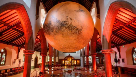 The sculpture, which is a large orange sphere, suspended from the cathedral roof. There are tables laid out underneath and the rafters are lit in red.