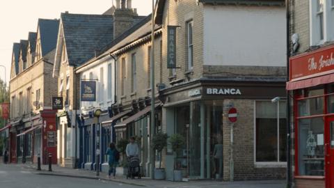 A street scene on a sunny day with of Walton Street's businesses - The Jericho Cafe, Branca restaurant, Jamais Indian Restautant and Daisies Flower Shop. A man is pushing a stroller on the street and a woman is walking past the Branca restaurant 