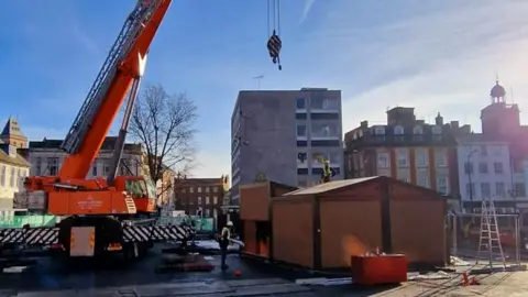 West Northamptonshire Council Orange crane above brown stall building in Market Square