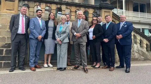 A crowd of people gathered infront of stone steps to a county hall wearing suits 