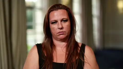 A woman wearing a black tank top and long reddish brown hair sits and stares at the camera.