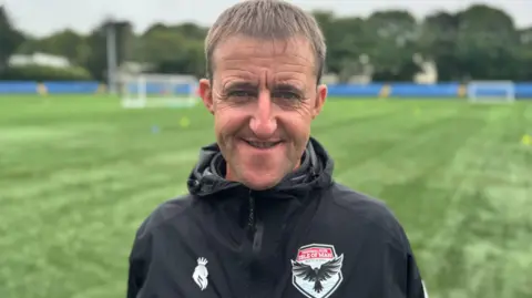 Lee Dixon wears a black rain jacket with the FC Isle of Man logo on the left side of his chest. He smiles. He has short fair hair and stands by a green football pitch.